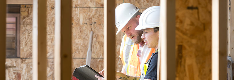 Two construction men with hardhats looking at an iPad inside a framed building.